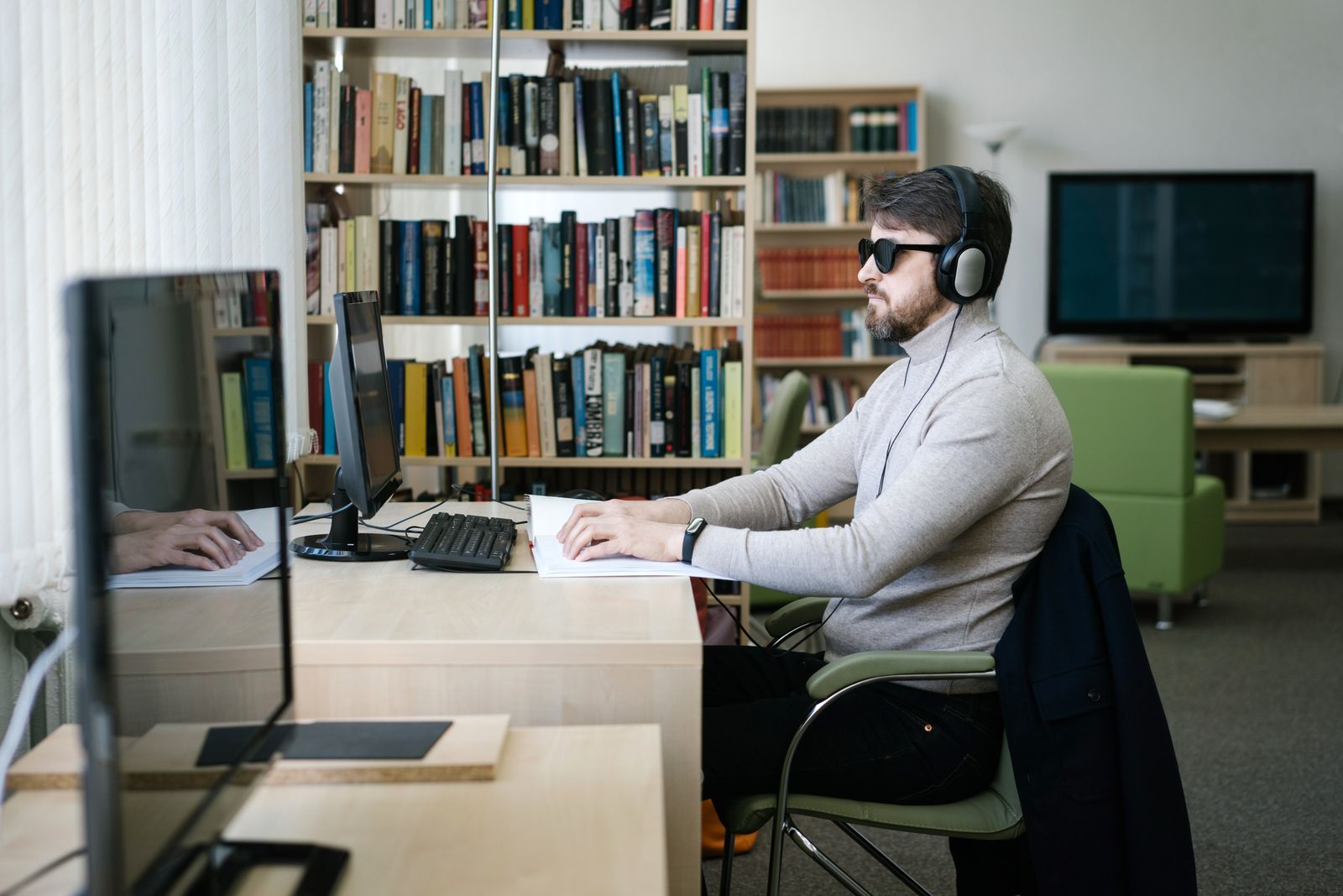 A blind man wearing headphones works independently at a computer in a library-style workspace.